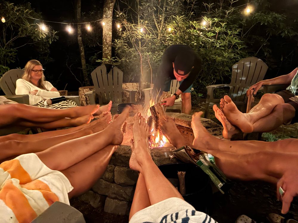 Bare feet stretched toward a stone fire pit at night, string lights overhead, while someone adds a log to the fire.