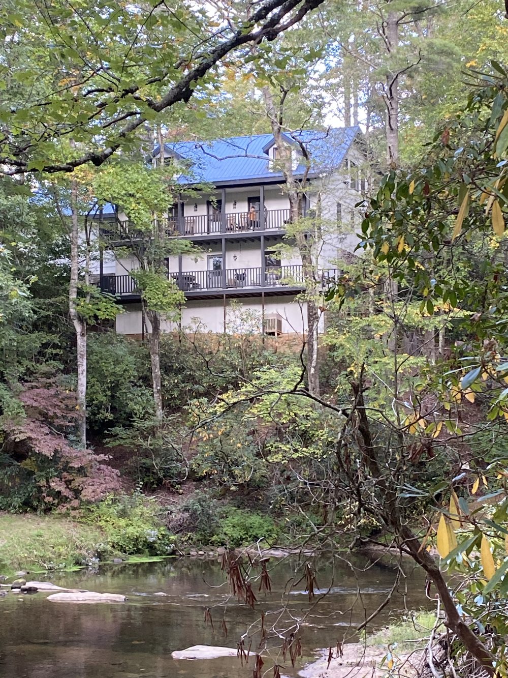 A three-story house with a blue metal roof and deep porches, set among trees on a hillside above a shallow rocky river.